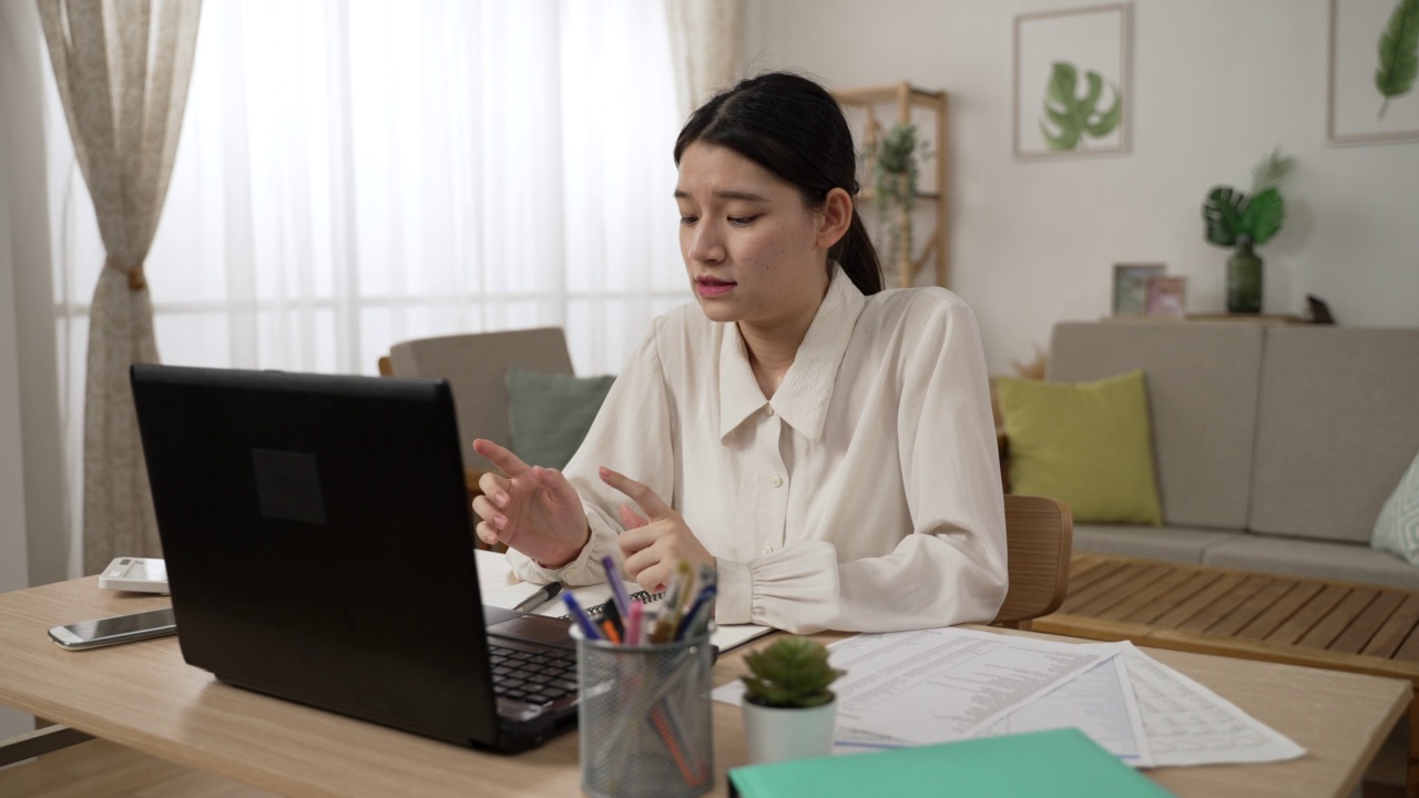 在家办公的亚洲女性商务人士在客厅里进行电话会议时，一边看着笔记本电脑屏幕，一边用手势交谈视频素材