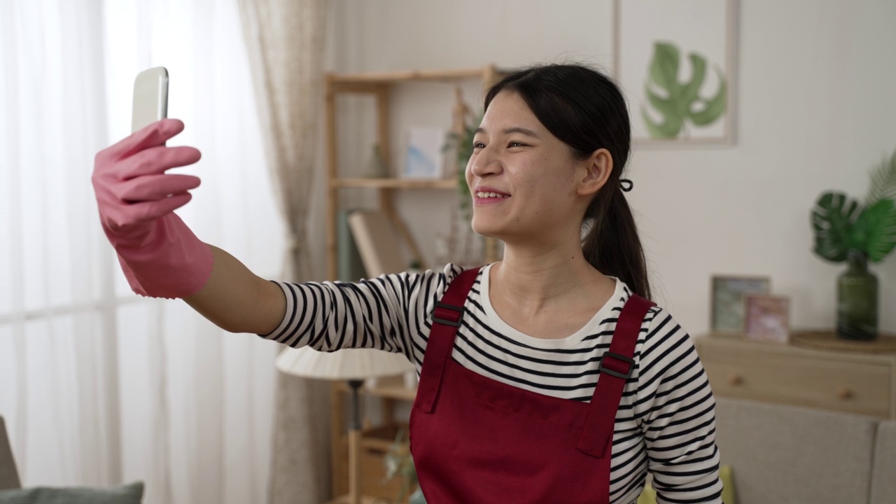 快乐的亚洲女孩在家庭围裙和橡胶手套是在电话屏幕和挥手，而与朋友视频电话在家里的客厅视频素材