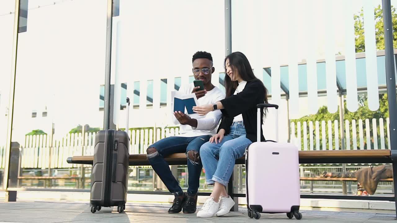 英俊微笑的年轻非洲裔美国人坐在电车站与他美丽的亚洲女性朋友和他们的护照照片，而等待在电车上视频素材