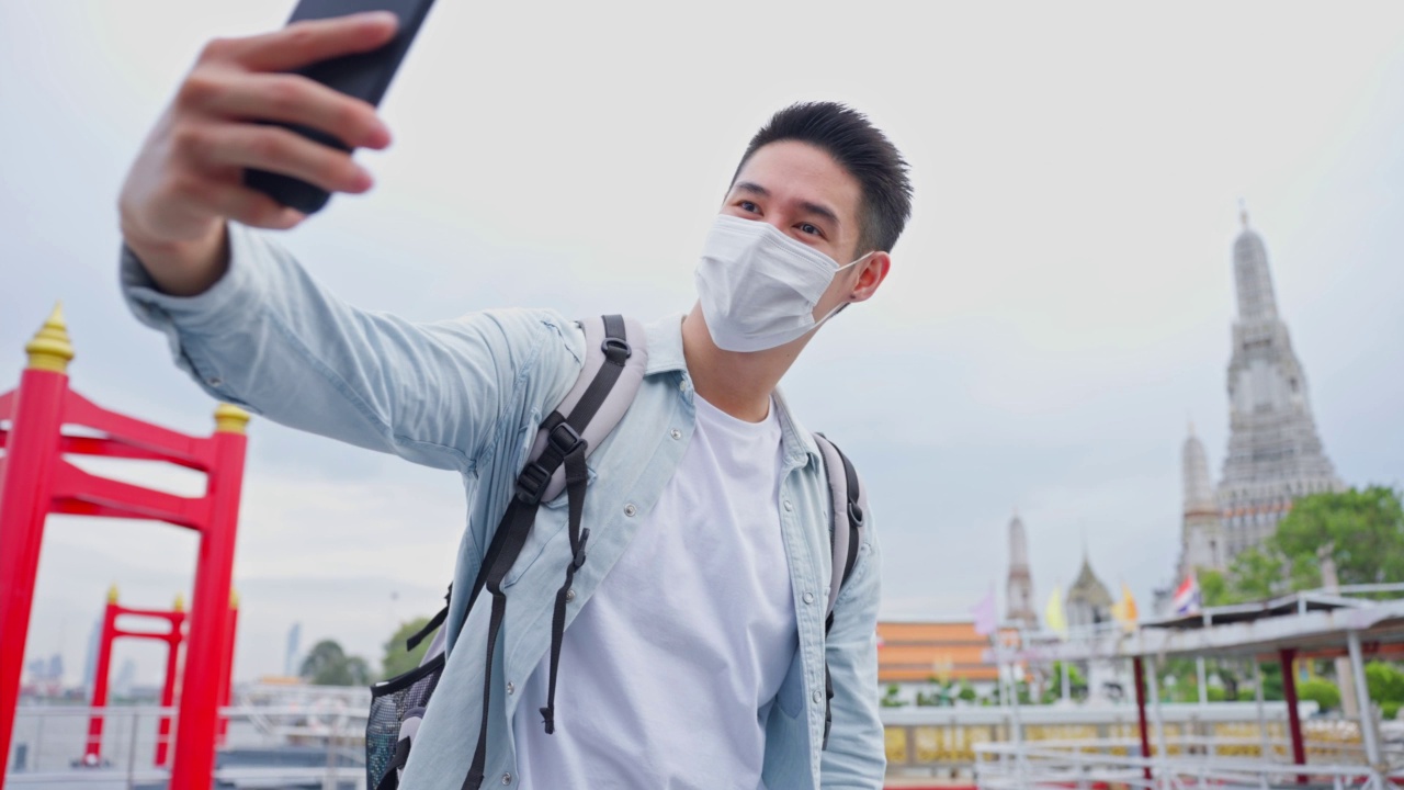 亚洲年轻男性背包客在城市里用智能手机自拍。帅哥旅游旅行者戴着口罩独自在街上旅行，用智能手机录视频，在泰国度假旅行视频素材