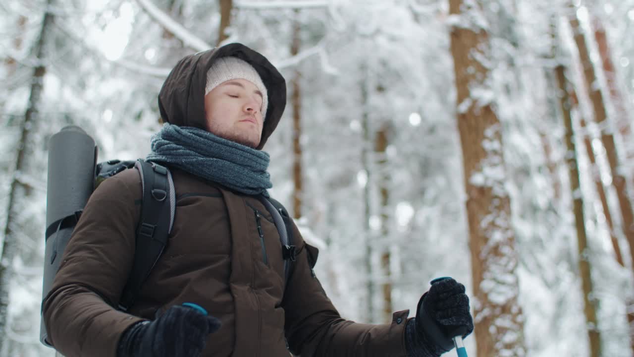 英俊的男性旅行者，带着登山杖和旅行背包站在冬天的森林里。年轻的白人徒步旅行者看着相机享受在山林中美妙的散步。在艰苦跋涉中休息视频素材