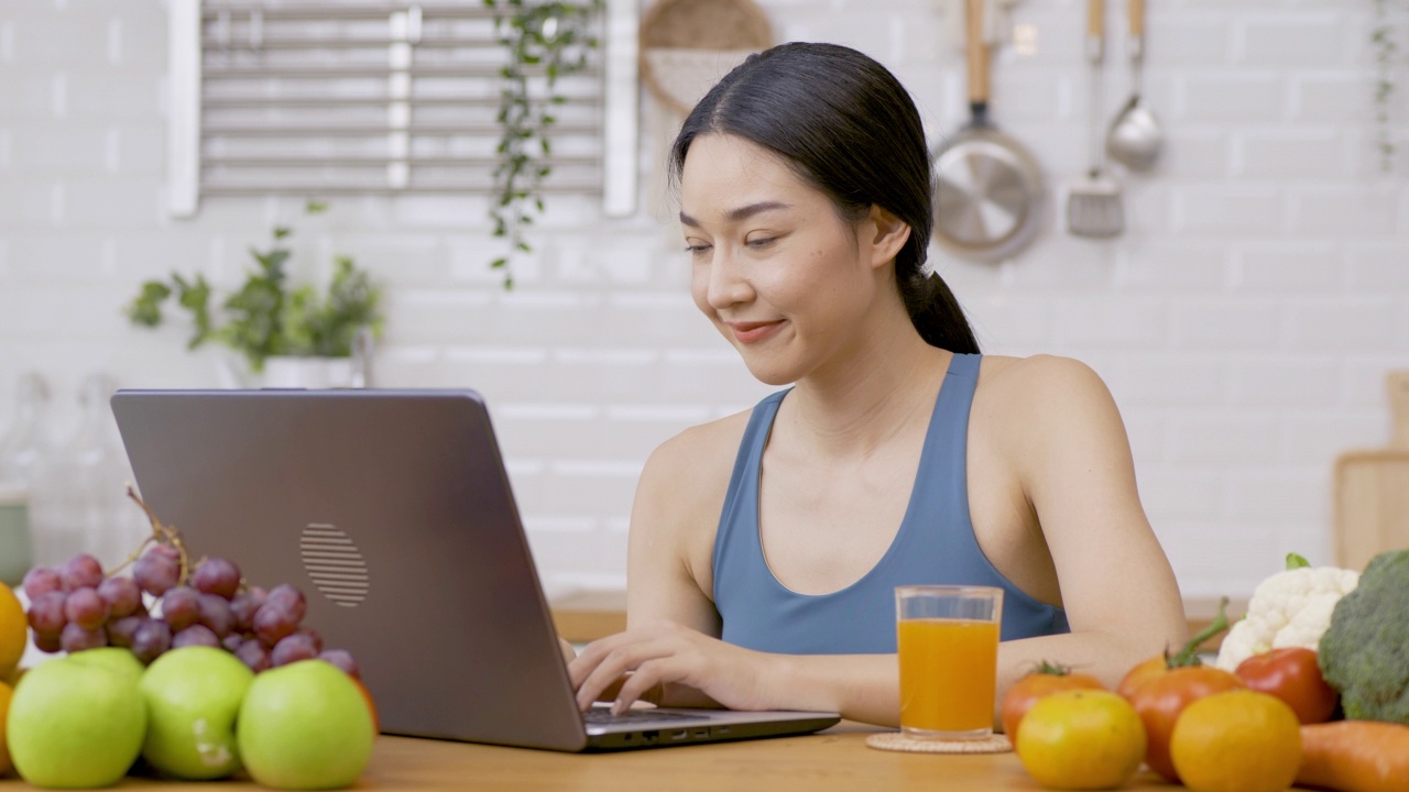 美丽的亚洲女人在厨房在家里。她在厨房用笔记本电脑，喝橙汁。保健和饮食的概念。视频素材