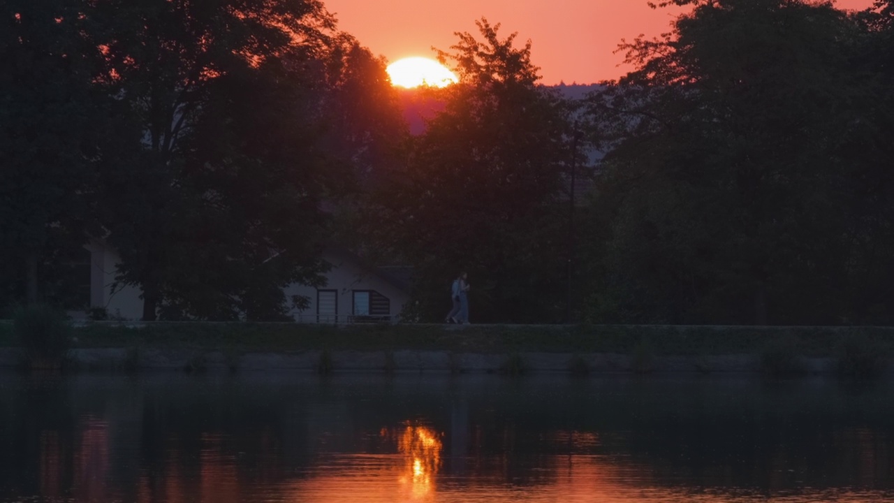 湖滨景观，在明亮的夕阳下，公园的树木倒映在湖水中，远处的行人在堤岸上行走视频素材