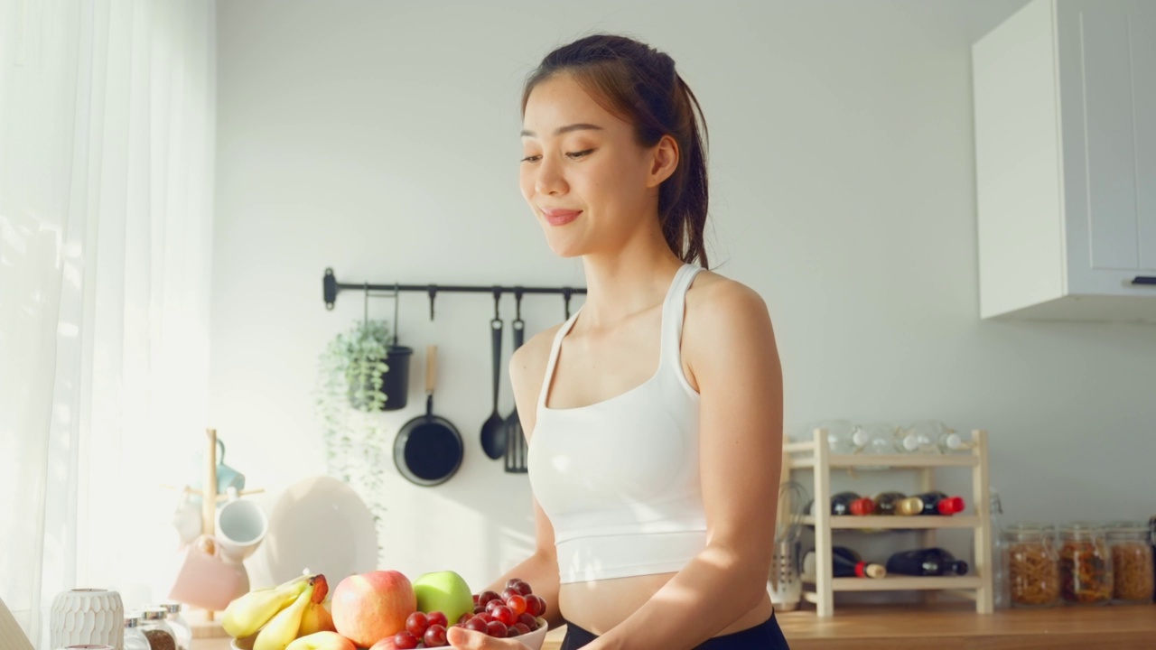 亚洲有吸引力的女人的肖像持有水果碗，看着相机。美丽的运动女孩在运动服享受吃干净的蔬菜锻炼后的健康在家里。饮食与健康食品理念。视频素材