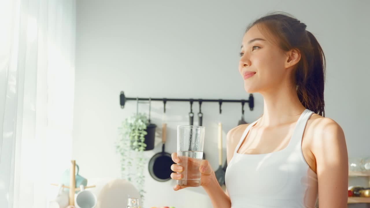 亚洲美女穿着运动服在家运动后喝水。年轻的口渴活跃的运动女孩喝干净的矿物自然在杯子锻炼后保健在厨房在房子。视频素材