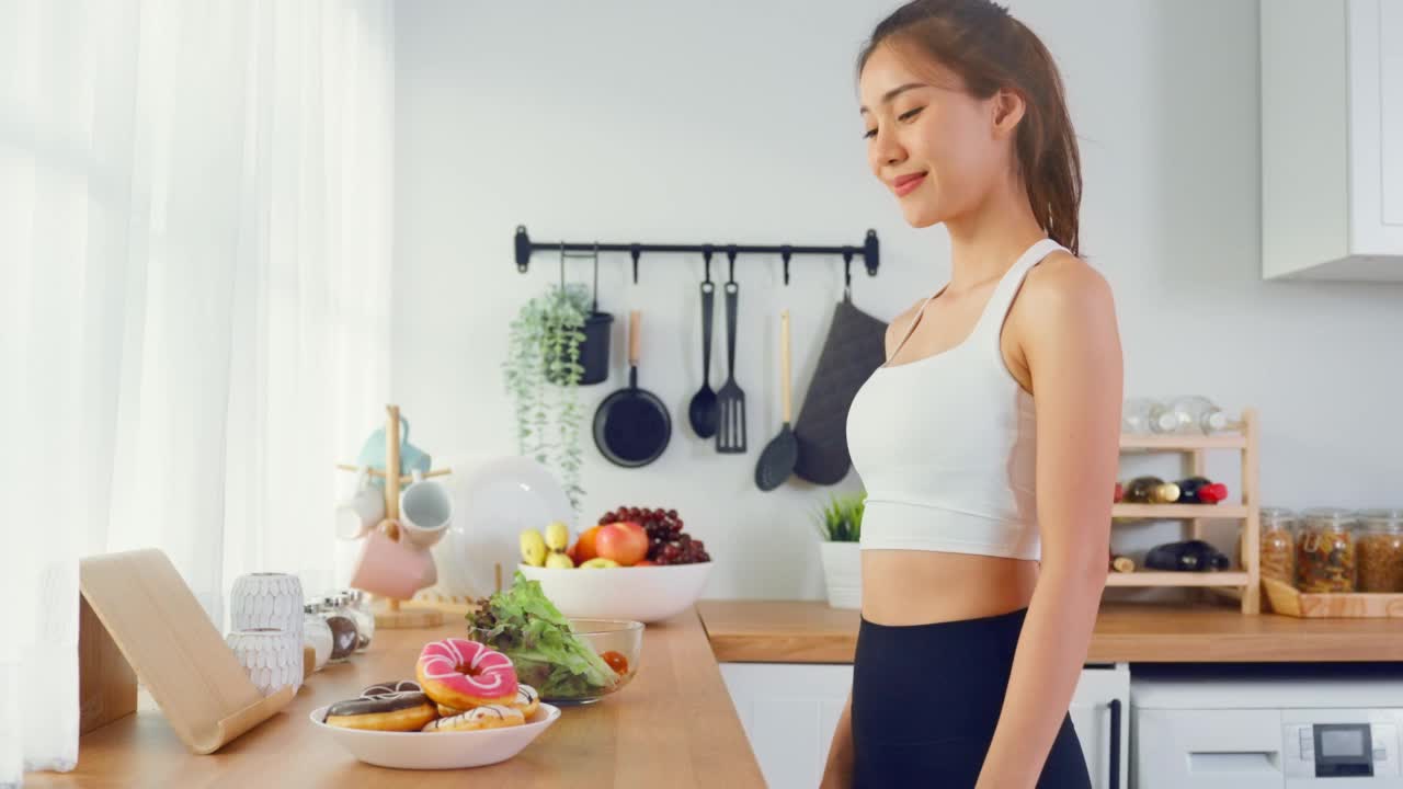 亚洲有吸引力的积极的女人拿起一个绿色沙拉在厨房在家里。漂亮的运动装女孩渴望选择水果，忽略甜甜甜圈，享受运动后吃健康食品的健康视频素材