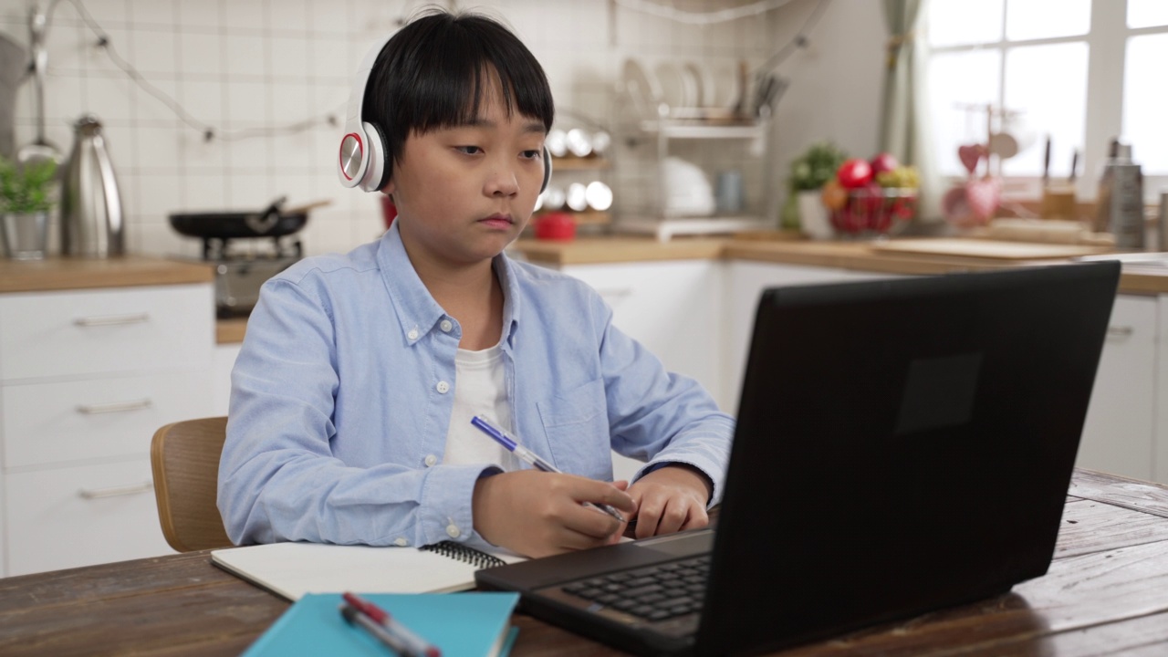 图为，一名亚洲学生戴着耳机，在餐桌上用笔记本电脑上在线课程。在家学习时，她一边点头，一边在笔记本上写字视频素材