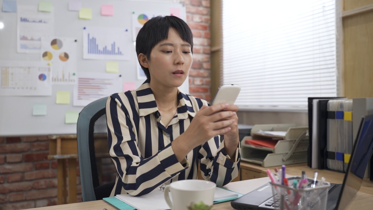 背景是一间明亮的阁楼办公室，一名认真的亚洲女员工正在办公桌上用智能手机发短信。视频素材