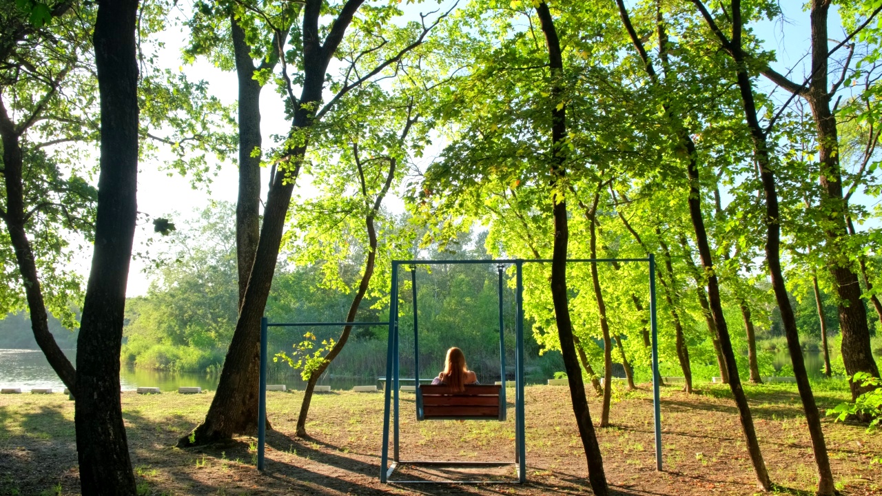 在金色夏日夕阳下荡秋千的女人。快乐的小女孩，戴着草帽，坐在秋千上，看着美丽的大自然视频素材