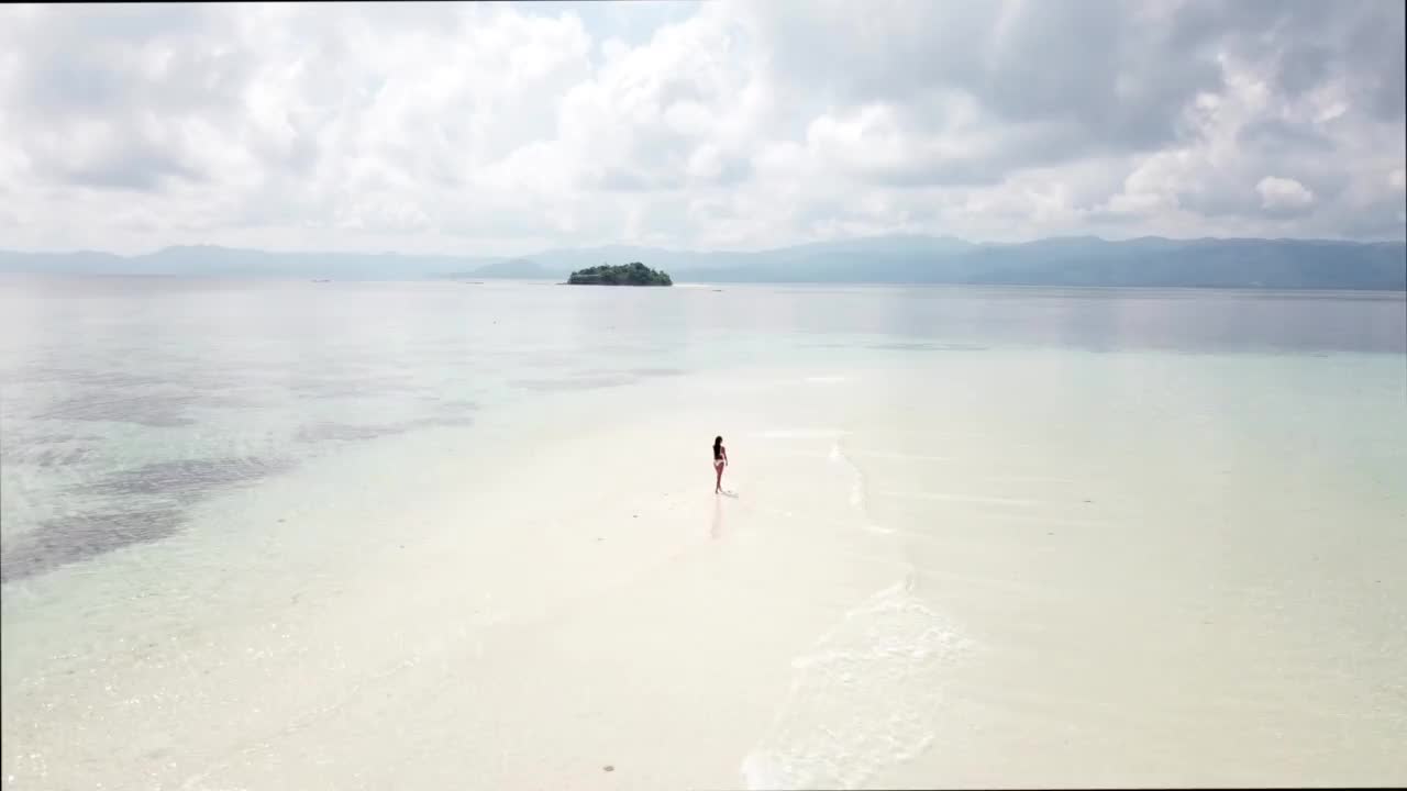 穿着比基尼的年轻女子走在白色的沙滩上，附近的蓝色热带海洋的波浪。视频素材