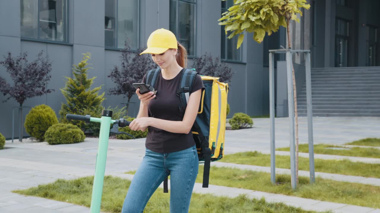 在办公室门口，一个骑着电动滑板车的送货女工在看电话。快递员背着背包，靠在电动滑板车的把手上看智能手机视频素材