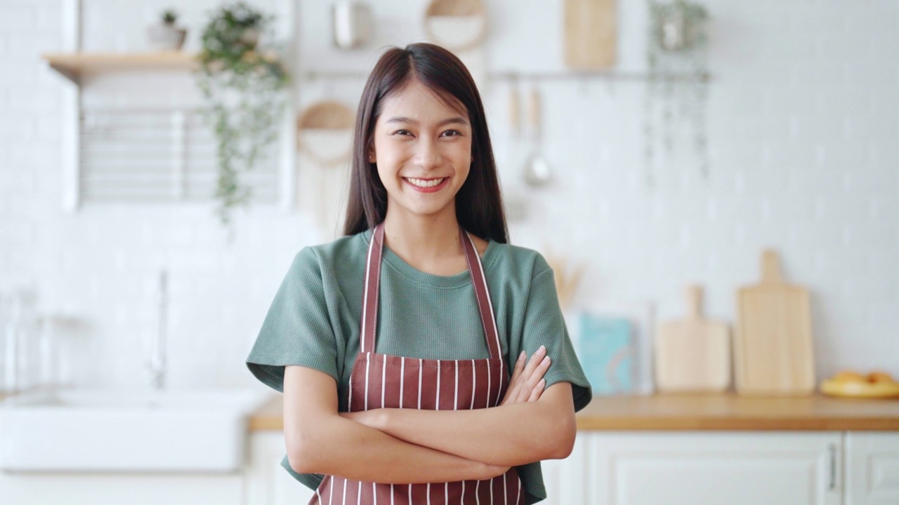 快乐的年轻亚洲妇女穿着围裙，站在厨房里。美丽的女性微笑着看着镜头感到自信视频素材