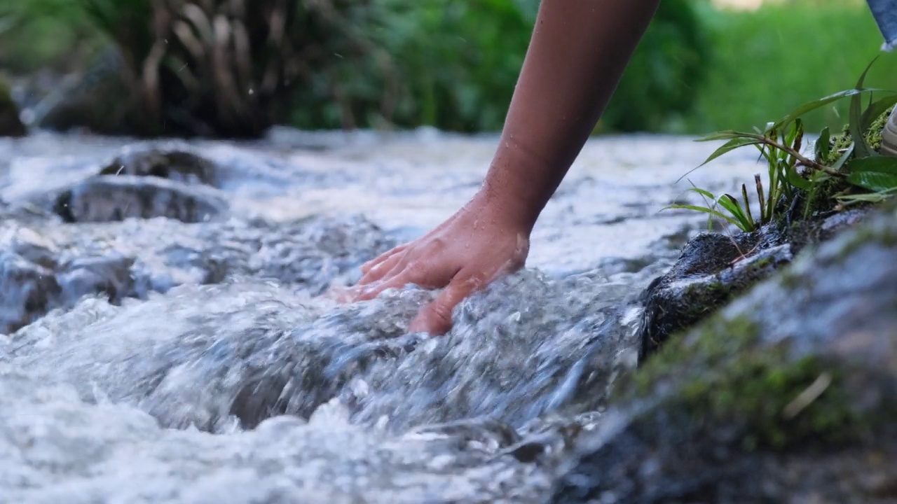 女人的手摸着冰冷的河水。一个徒步旅行的女人站在一块岩石上，用她的手触摸一条小溪。手的特写视频素材