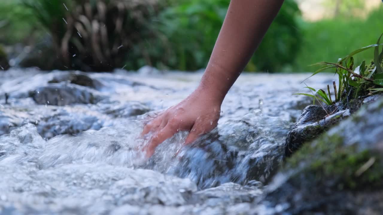 女人的手摸着冰冷的河水。一个徒步旅行的女人站在一块岩石上，用她的手触摸一条小溪。手的特写视频素材