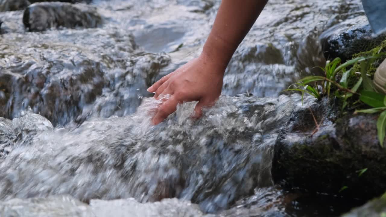 女人的手摸着冰冷的河水。一个徒步旅行的女人站在一块岩石上，用她的手触摸一条小溪。手的特写视频素材