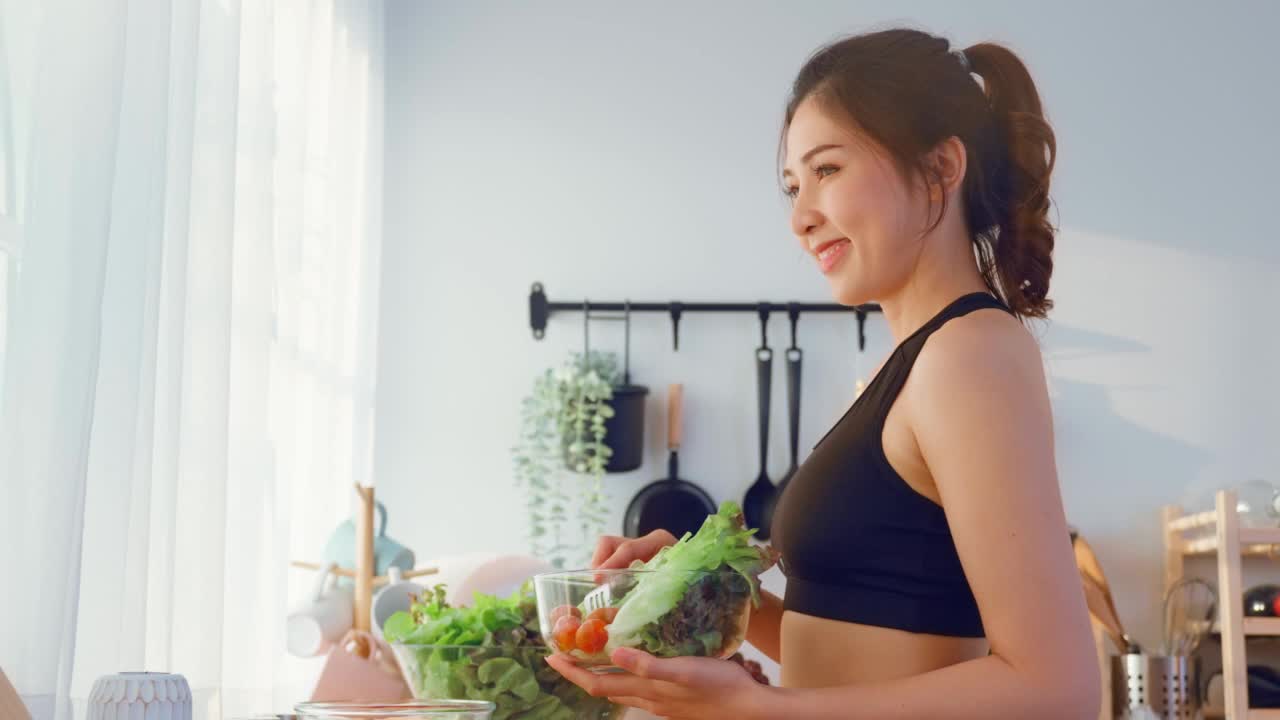 亚洲吸引人的运动女子拿着沙拉碗吃蔬菜。美丽的运动女孩在运动服享受吃干净的蔬菜锻炼后的健康在家里。饮食与健康食品理念。视频素材