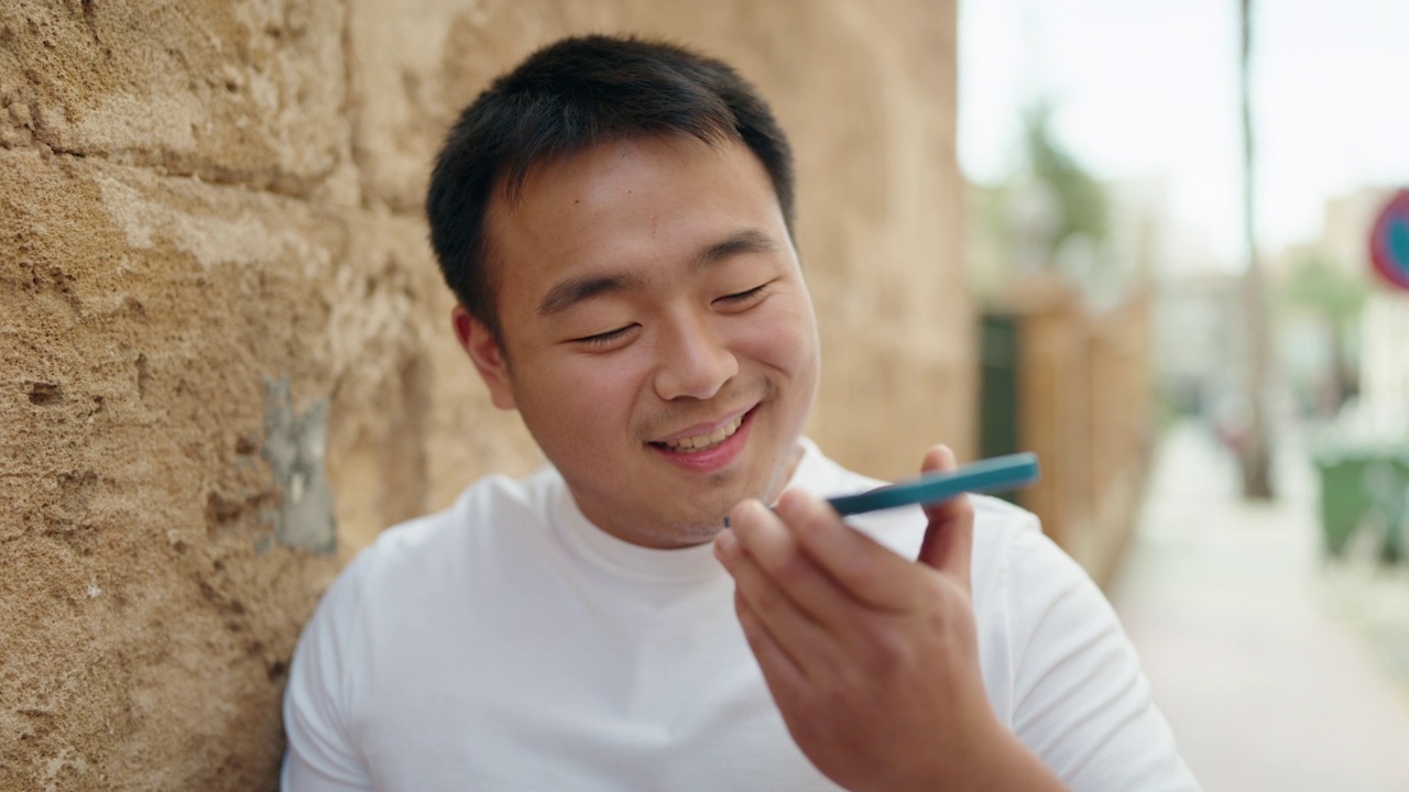 一名年轻的中国男子微笑着，自信地在街上用智能手机聊天视频素材