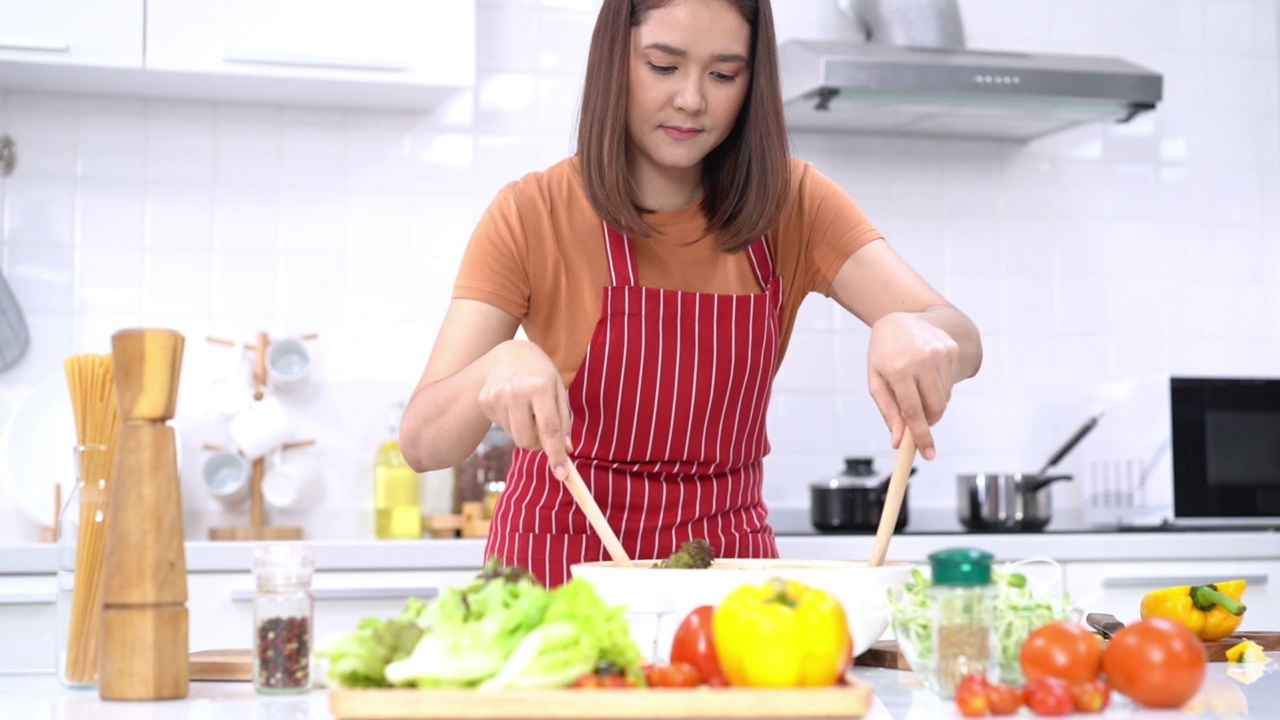 健康的生活方式和饮食观念。年轻的亚洲妇女准备沙拉在家里厨房。快乐微笑美丽的年轻女子准备健康的素食沙拉在家里厨房。视频素材