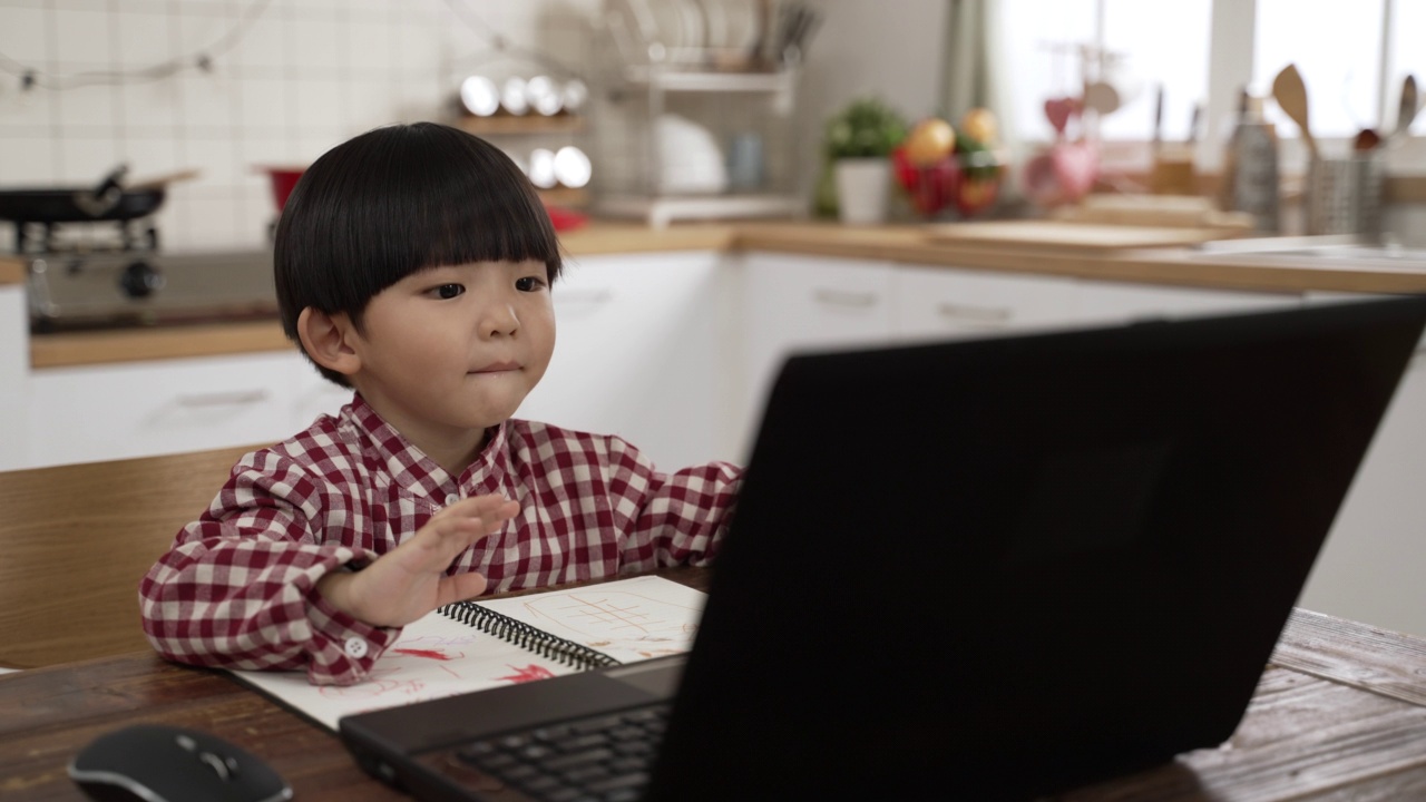 可爱的亚洲男学生在家里的餐厅上在线课程，对着笔记本电脑屏幕挥手打招呼。在家学习的概念视频素材