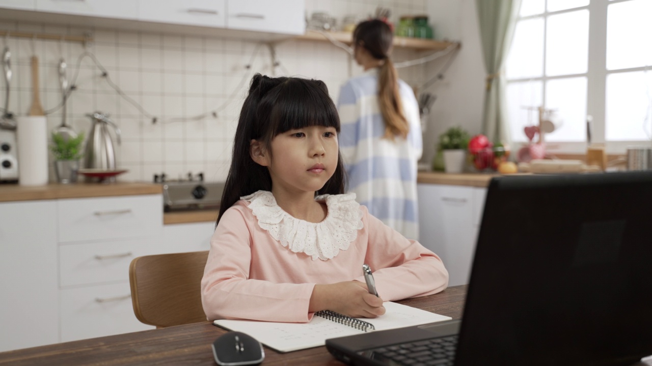 在笔记本电脑上观看在线学习视频时，专注的亚洲女孩点头的慢动作。她写作业时，妈妈在厨房里做饭视频素材