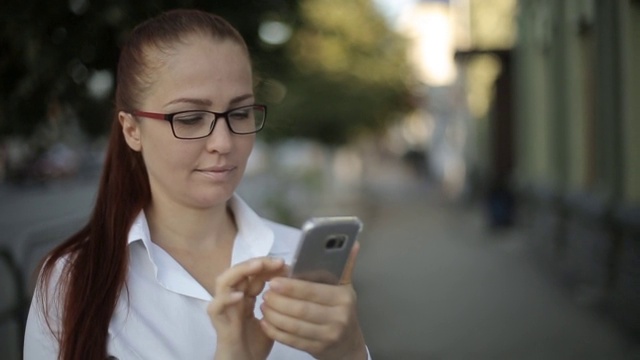 一头红头发、戴着眼镜、穿着白色上衣的白人中年女性正在通过智能手机屏幕查看电子邮件。视频素材