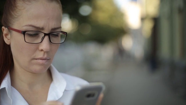 一头红头发、戴着眼镜、穿着白色上衣的白人中年女性正在通过智能手机屏幕查看电子邮件。视频素材