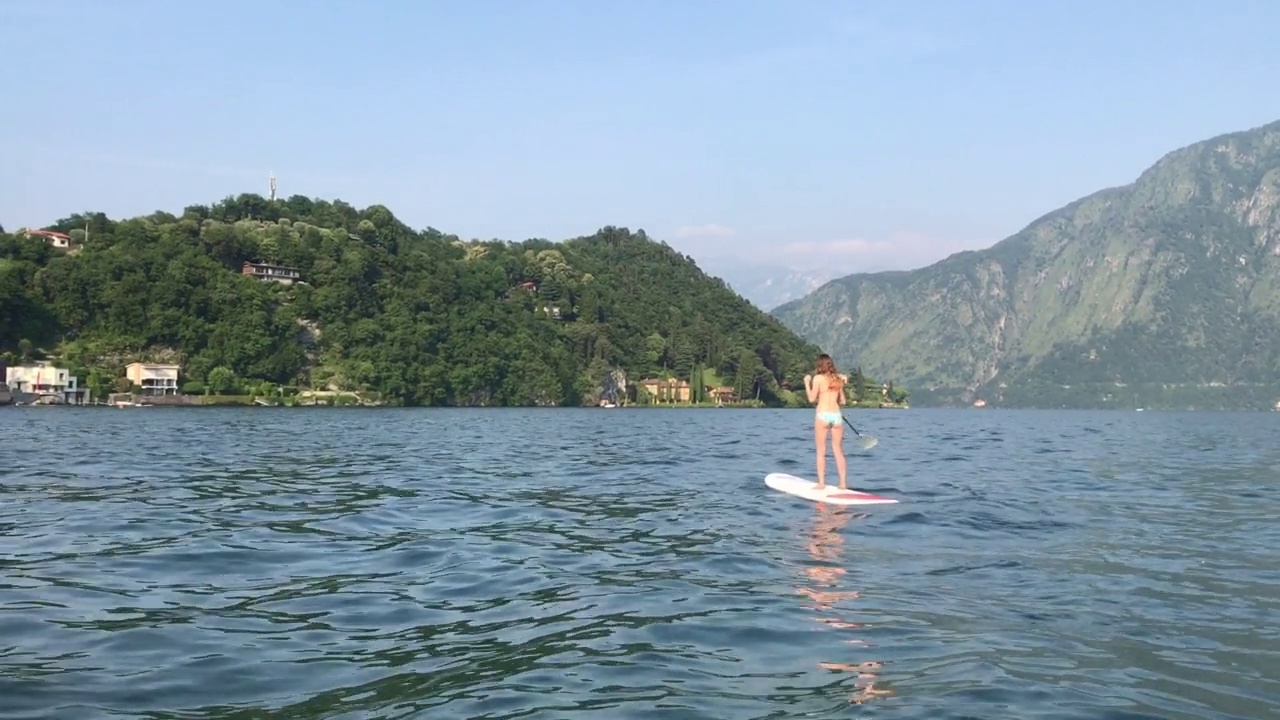 在阳光明媚的日子里，年轻的女子在山上的湖上站着划桨板视频素材