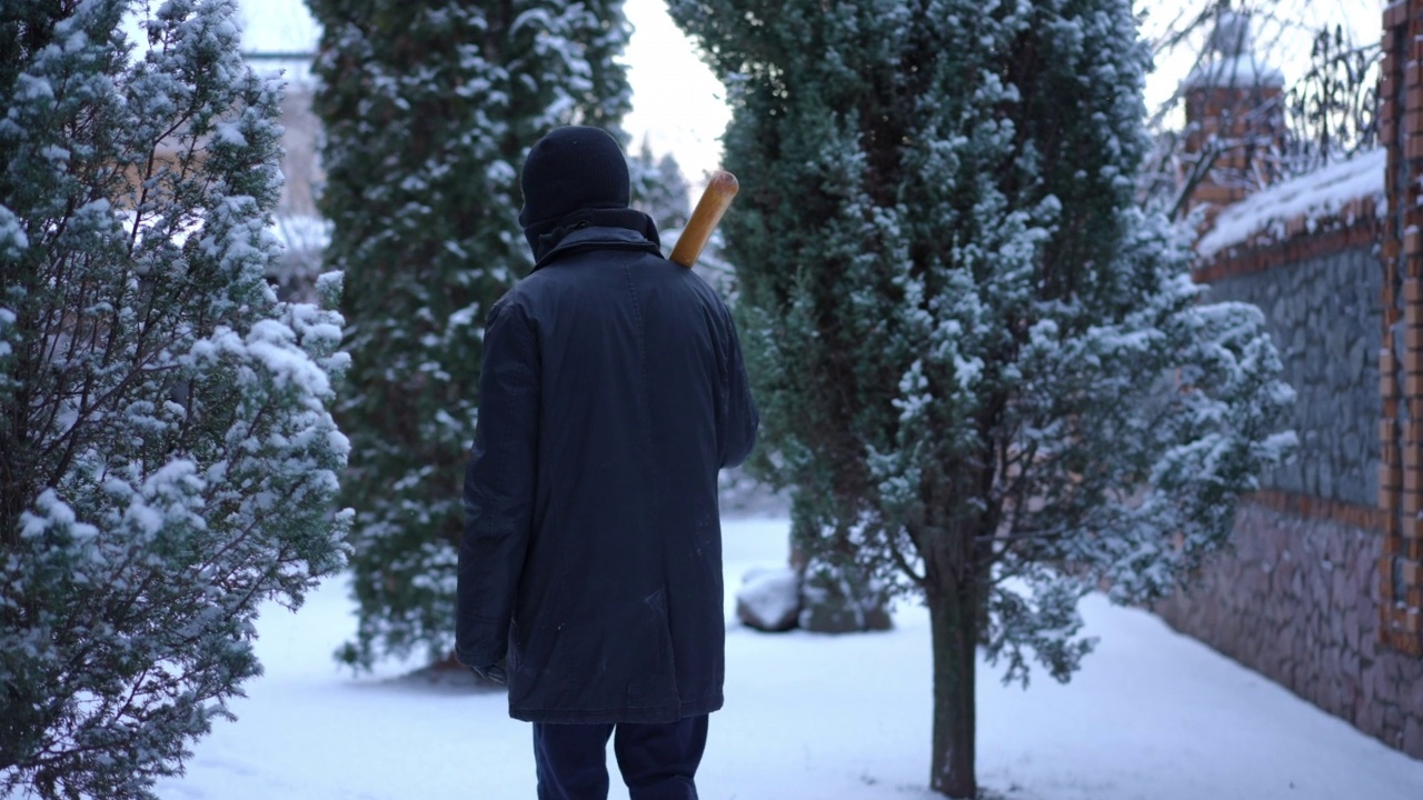 带着棒球棒的夜贼走在外面积雪的后院。现场摄像机记录了一名戴着滑雪面罩的自信男子在白雪上漫步，四处张望。视频素材