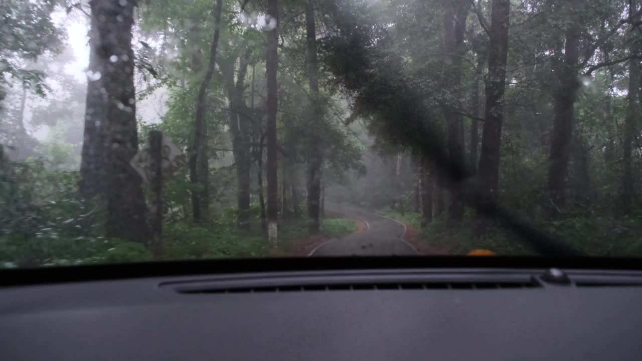 在雨中驾驶时，车内的雨点落在挡风玻璃上，雨刷也在工作。在一个下雨天，在森林里的路上开车。视频素材