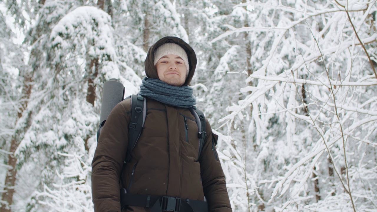 肖像年轻愉快的旅行者徒步旅行背包放松在冬天的森林降雪。快乐的人看着镜头，微笑着享受在树林里的跋涉。雪花纷纷落下。人与自然视频素材