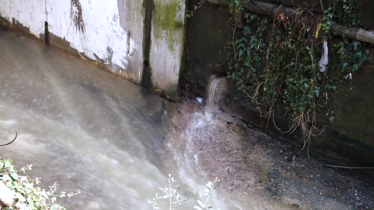 下水道从墙上的一个洞流出来，流入下水道。一股雨水从长满苔藓的水泥墙上的一个洞里流进一条小溪。视频下载