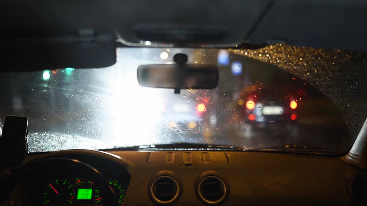 一个人在黑暗的雨城中穿过一场暴雨。当一个人开车时，夜晚的灯光和雨会穿过车辆内部。玻璃上的雨滴视频素材