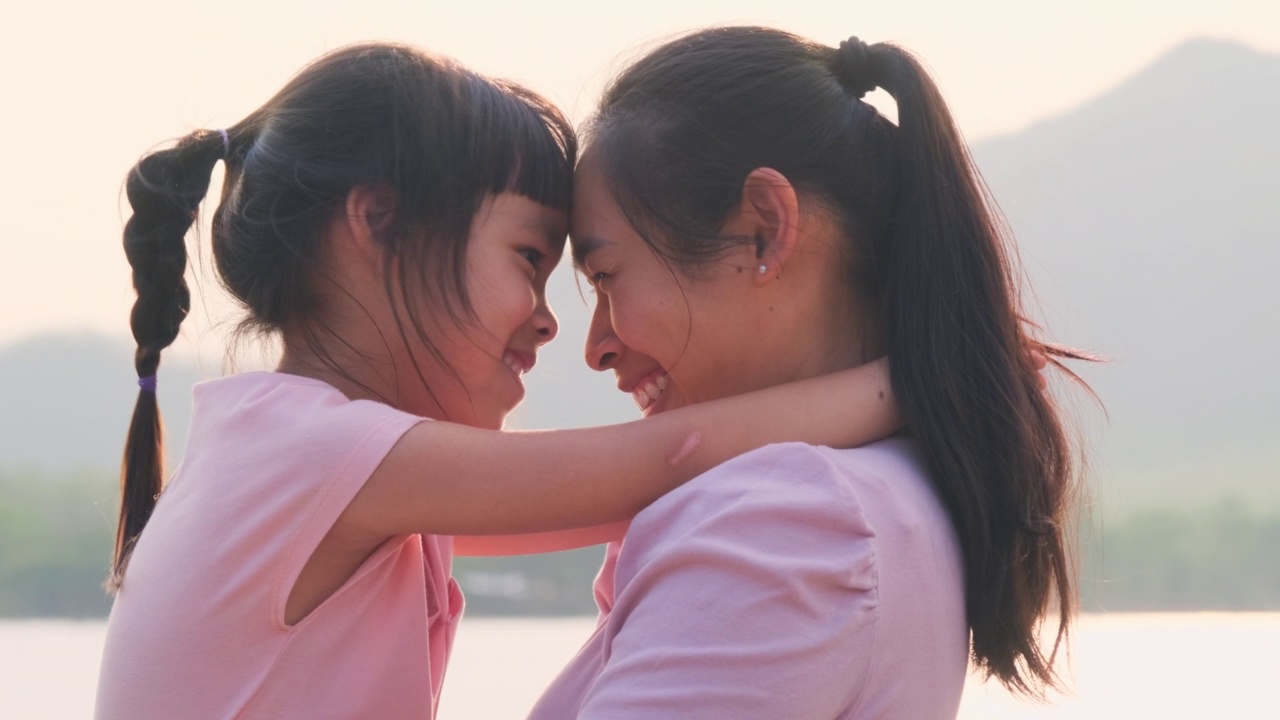 夕阳西下，母亲抱着可爱的小女儿在湖边散步。兴奋的小女孩笑着和妈妈在户外玩。母亲拥抱着女儿。视频素材