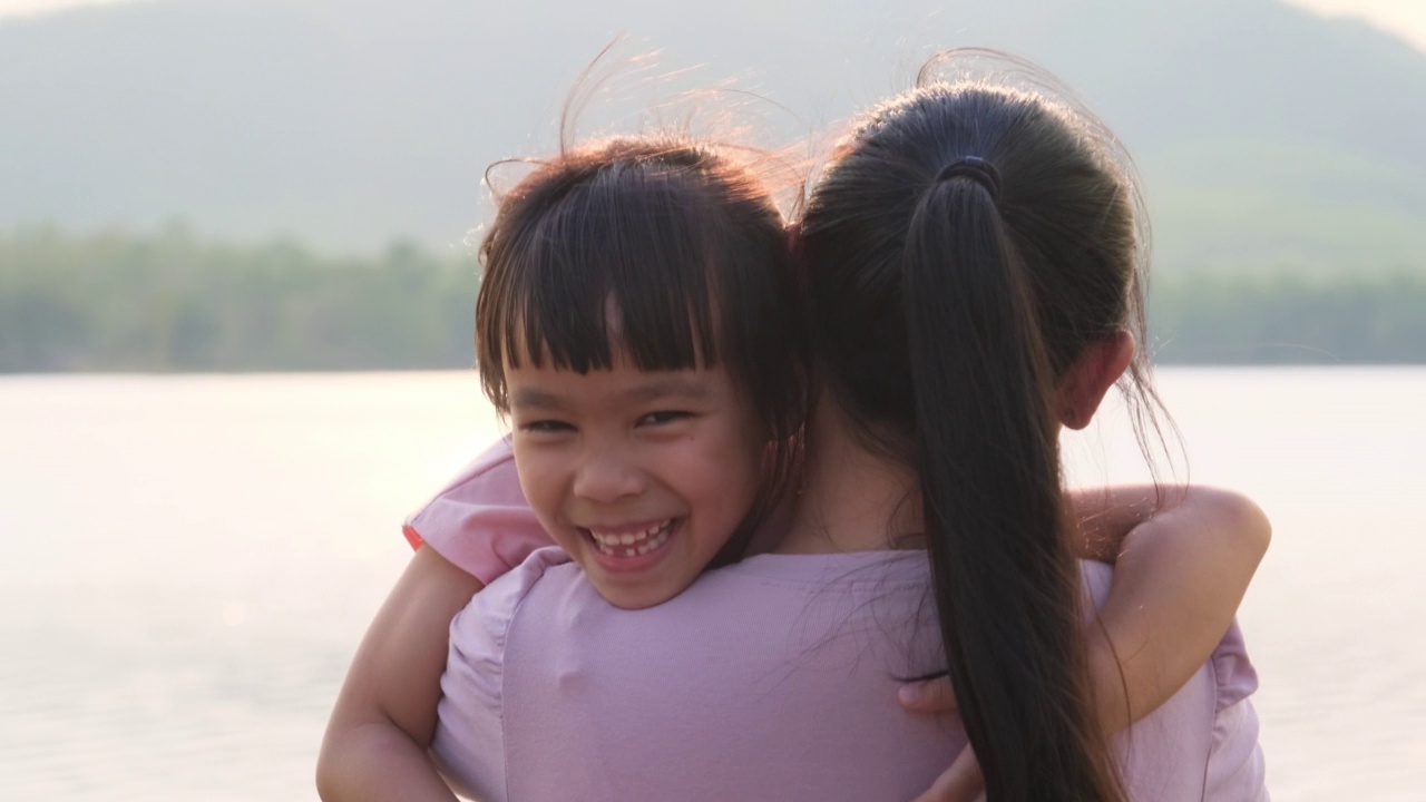 夕阳西下，母亲抱着可爱的小女儿在湖边散步。兴奋的小女孩笑着和妈妈在户外玩。母亲拥抱着女儿。视频素材