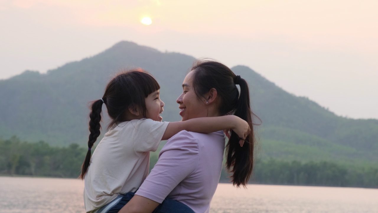 夕阳西下，母亲抱着可爱的小女儿在湖边散步。兴奋的小女孩笑着和妈妈在户外玩。母亲拥抱着女儿。视频素材