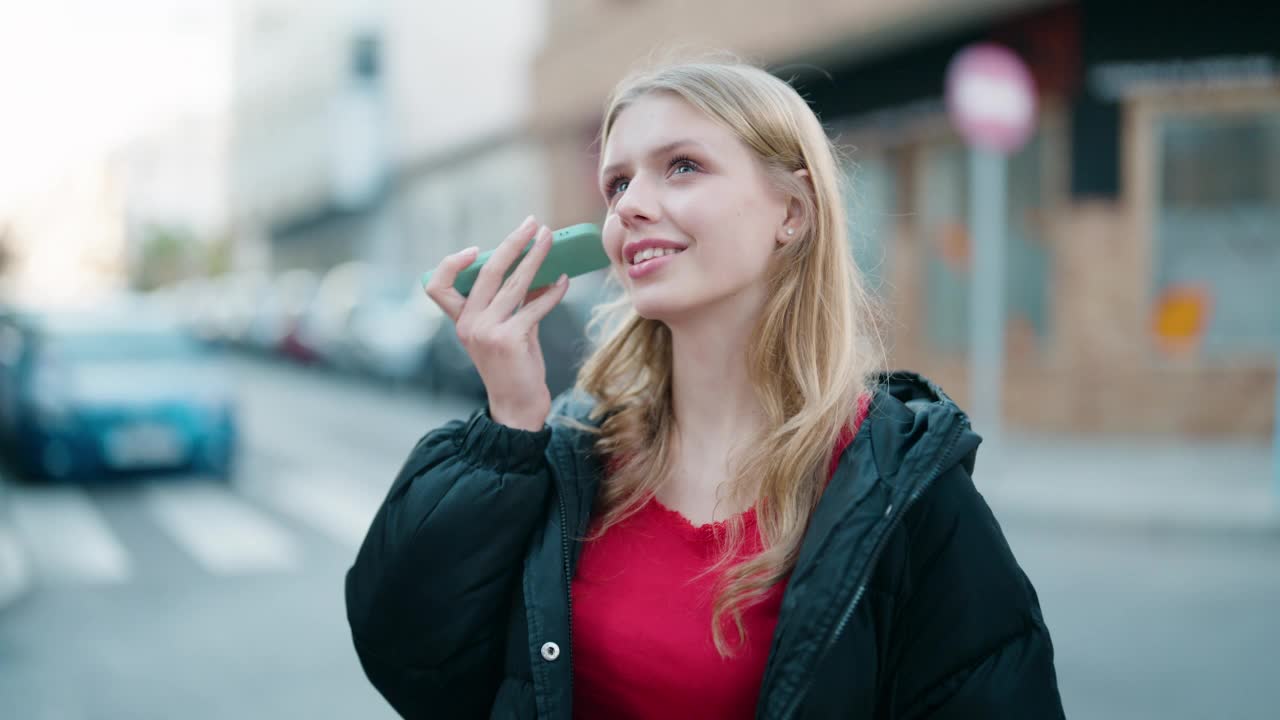 年轻的金发女孩微笑着自信地听着语音信息在街上的智能手机视频素材