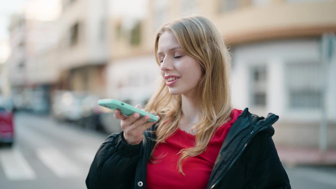 年轻的金发女孩微笑着自信地在街上对着智能手机说话视频素材