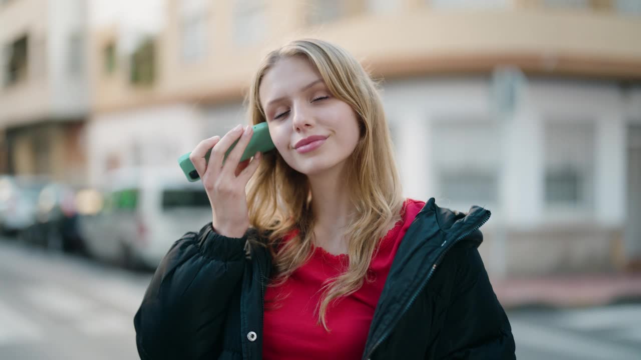 年轻的金发女孩微笑着自信地听着语音信息在街上的智能手机视频素材