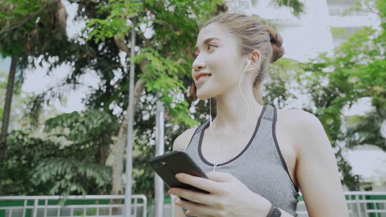 年轻美丽的亚洲女人听音乐从耳机慢跑锻炼在公园在夏天的早晨。健康的女运动员享受户外运动训练，在城市跑步锻炼视频素材