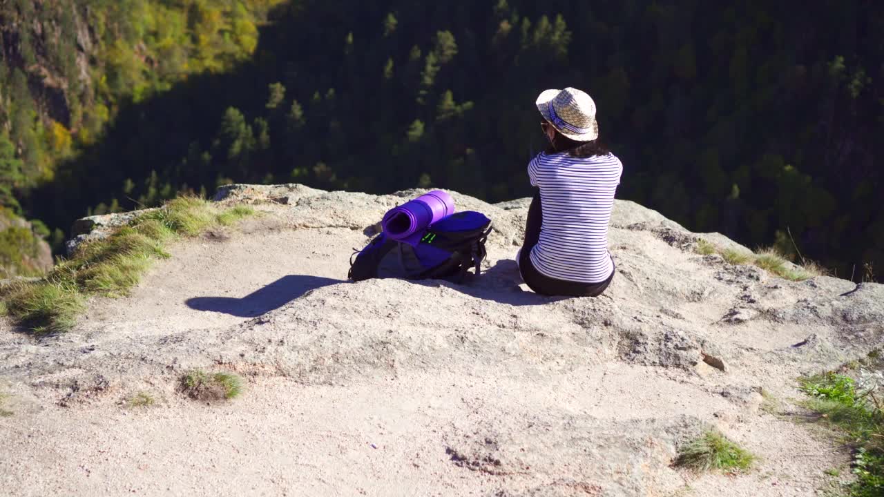 一名年轻的游客戴着帽子坐在山顶上，看着山上美丽的风景，躺在背包旁边，近距离观看视频素材