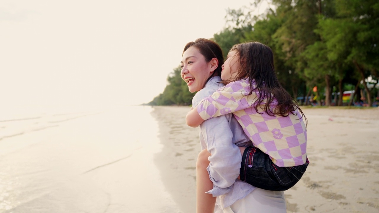 4K亚洲母亲背着小女儿在夏天的夕阳下漫步在热带海滩上。视频素材