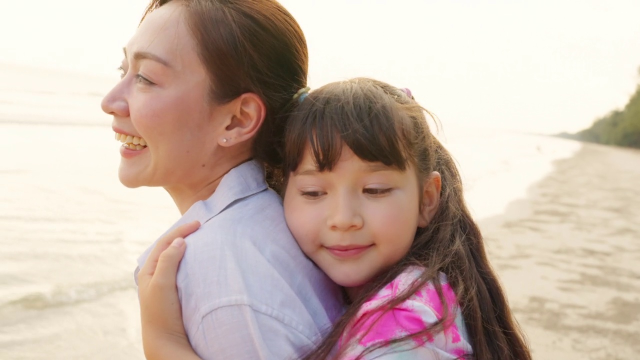 4K亚洲母亲背着小女儿在夏天的夕阳下漫步在热带海滩上。视频素材