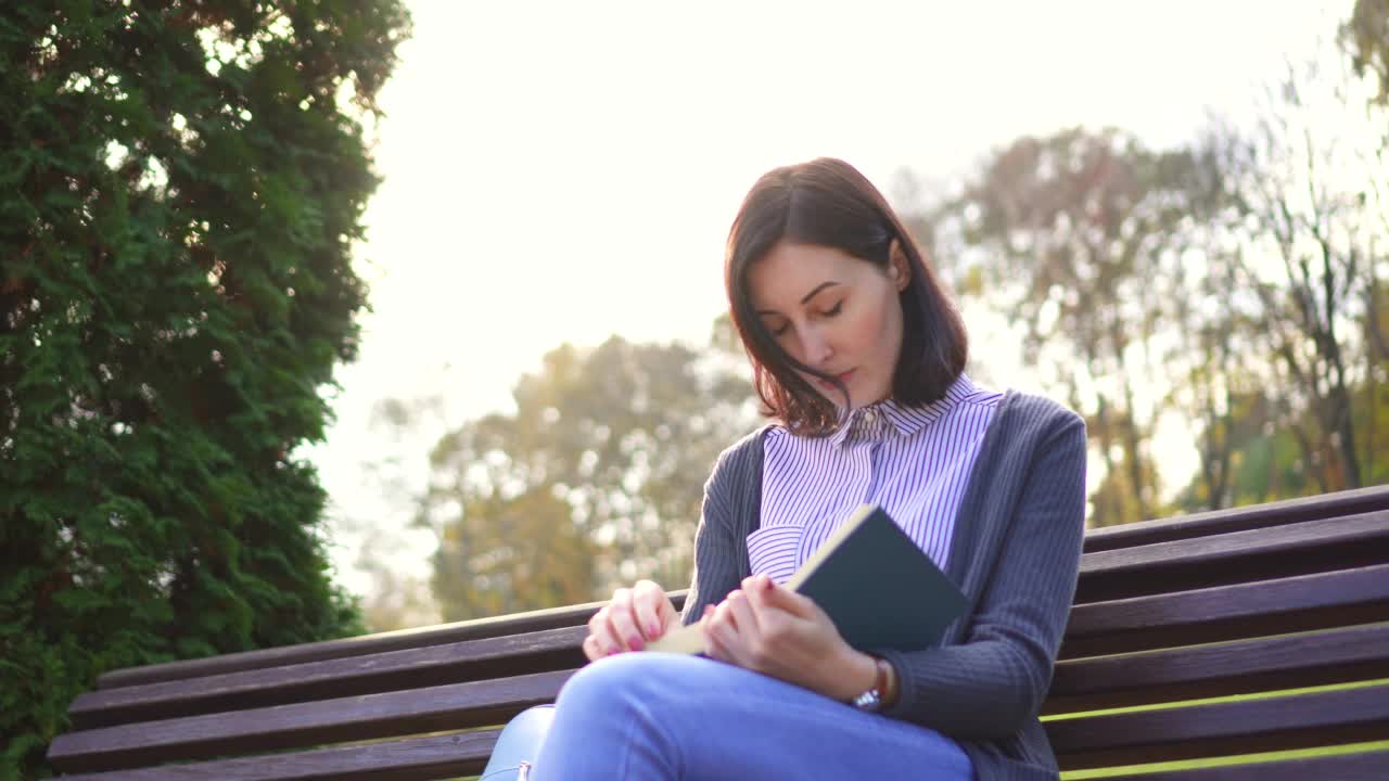 漂亮的女孩，膝盖上放着一本书，坐在户外的长椅上视频素材