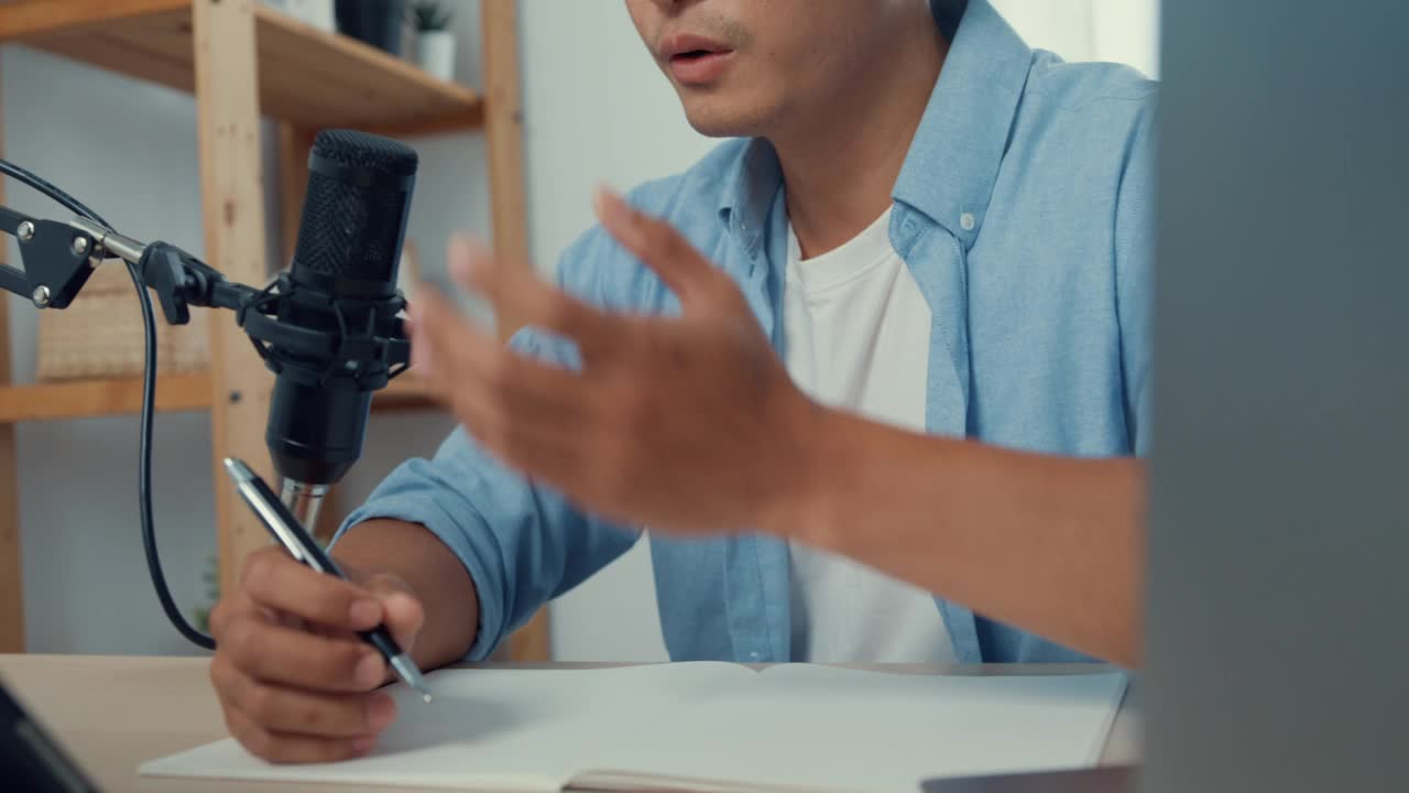 近距离的年轻亚洲男子录制播客在她的笔记本电脑与显微镜坐在家里客厅的办公桌上。视频素材