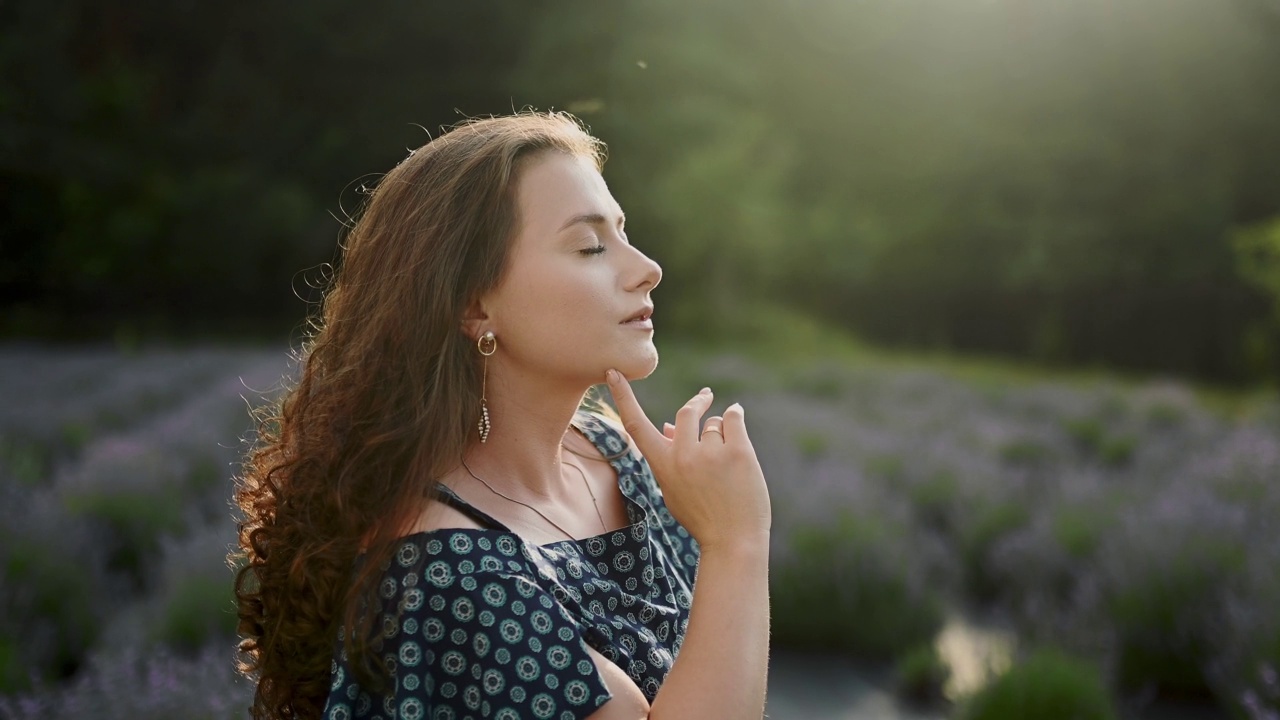 微笑的年轻白人黑发女人的肖像脸，站在薰衣草田，在相机摆姿势，阳光天气的风吹卷发，生活方式，户外，慢动作视频下载