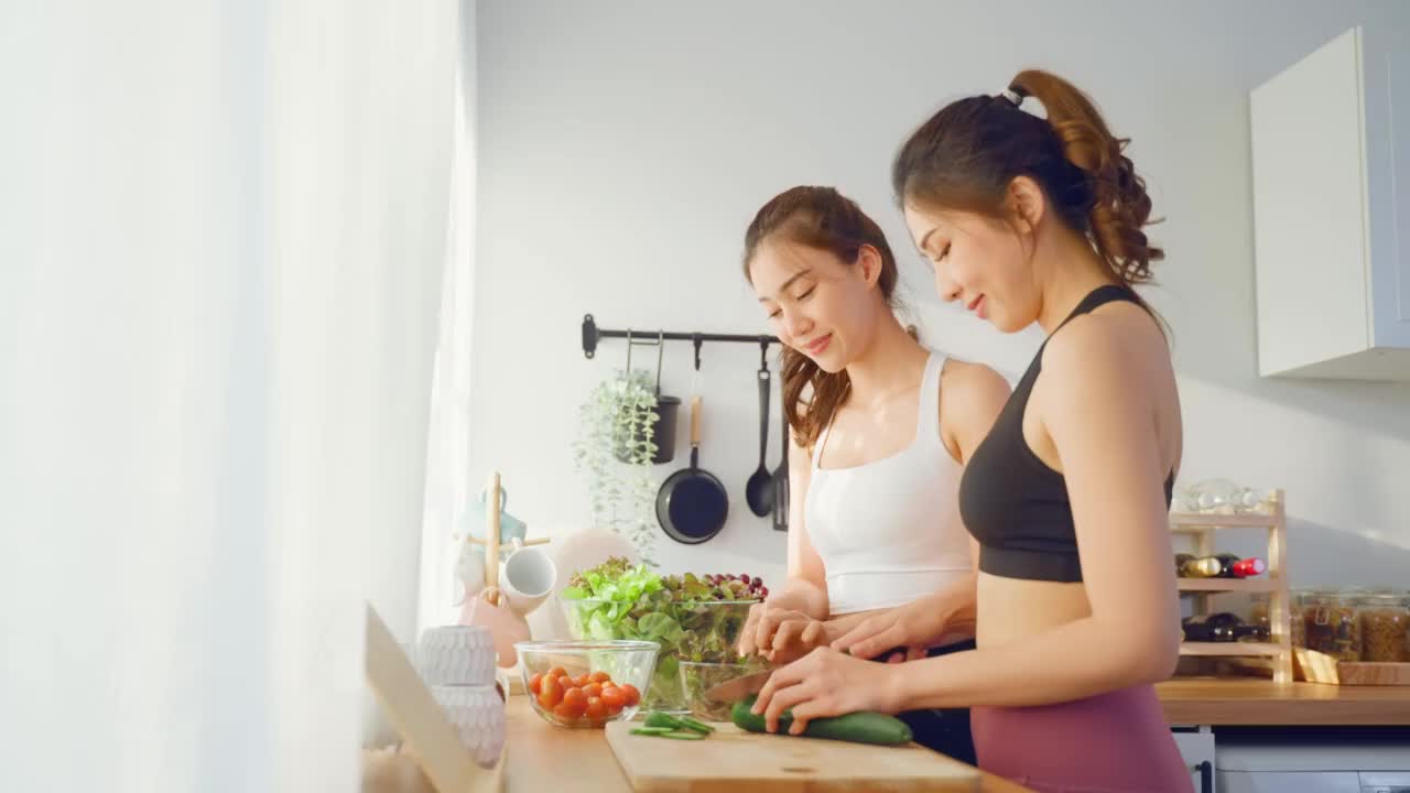 亚洲年轻的两个女子兄妹在厨房里穿着运动服烹饪沙拉。活泼漂亮的女孩妹妹心情愉快，喜欢吃蔬菜，健康饮食，减肥保健在家里视频素材