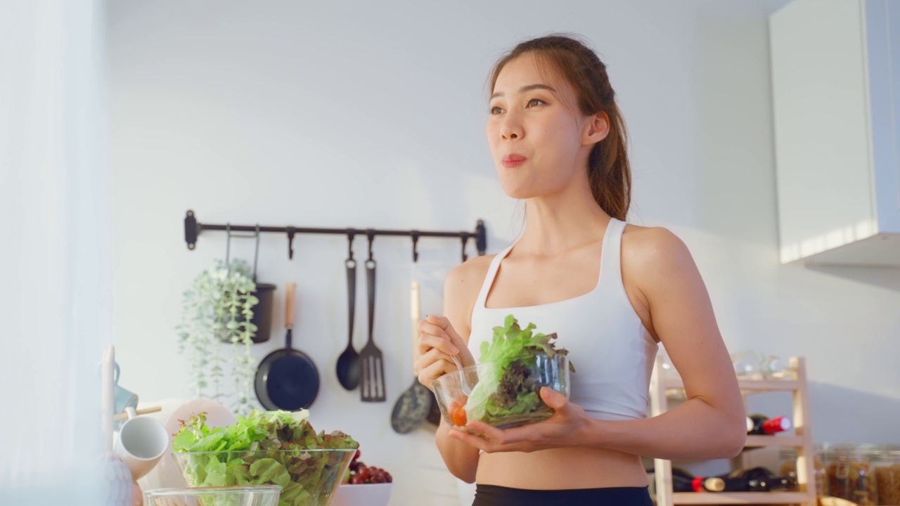 亚洲吸引人的运动女子拿着沙拉碗吃蔬菜。美丽的运动女孩在运动服享受吃干净的蔬菜锻炼后的健康在家里。饮食与健康食品理念。视频素材