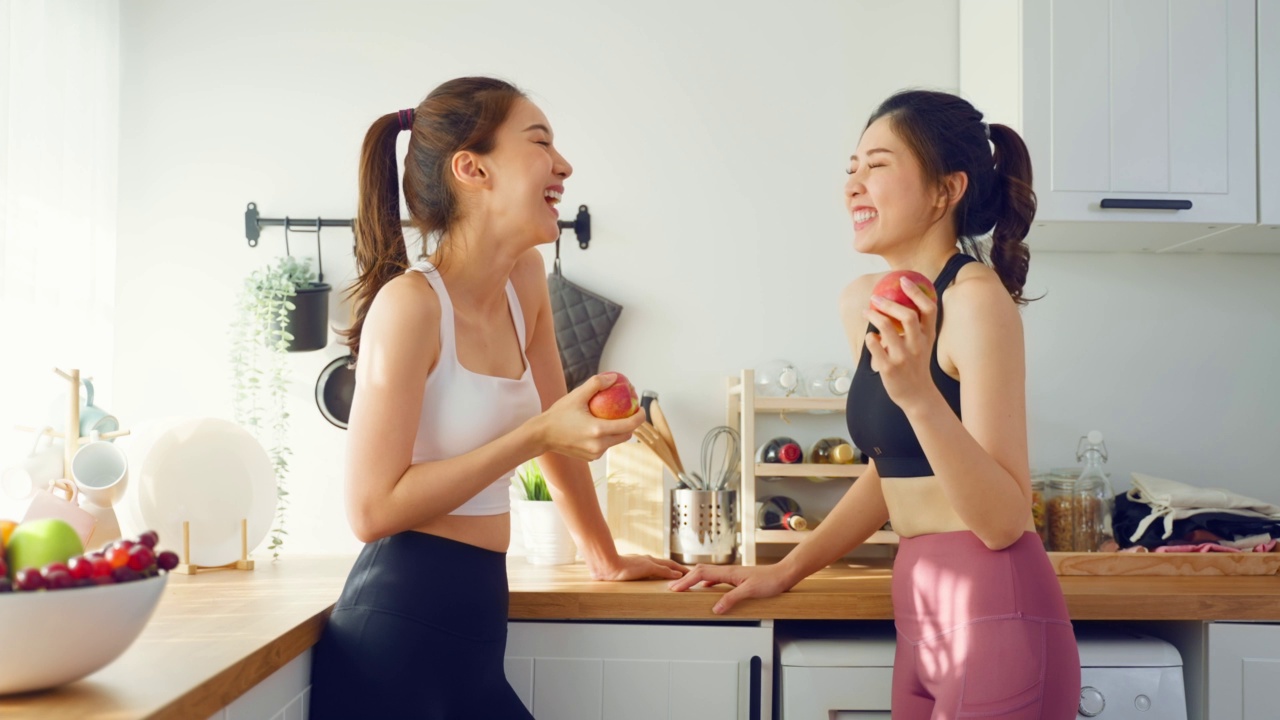 亚洲活跃的两个姐妹穿着运动服在厨房里吃苹果。年轻漂亮的女孩妹妹心情愉快，喜欢吃水果，健康食品，饮食和减肥保健在家里。视频素材