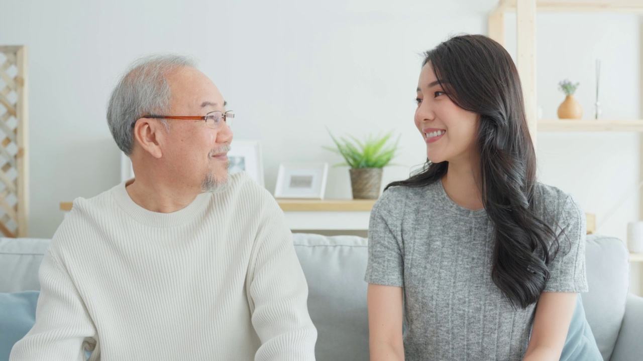 亚洲美女女儿的肖像拥抱和坐在年长的父亲。迷人的女人孙女拜访年长的成熟男人在家里，享受花时间一起在客厅看着相机视频素材