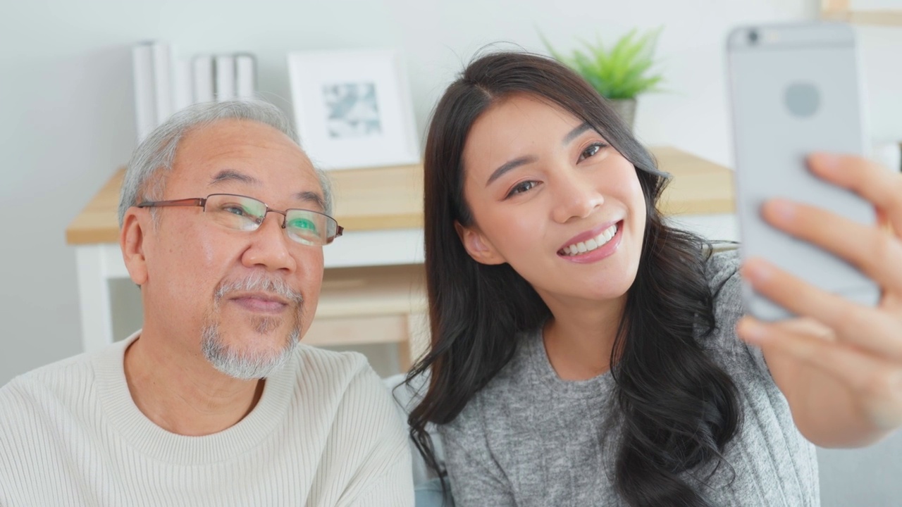 亚洲可爱的家庭，女儿用电话与资深父亲视频通话。年轻漂亮的女孩和上了年纪的老男人坐在沙发上享受网上交流，在家里的客厅里一起聊天视频素材